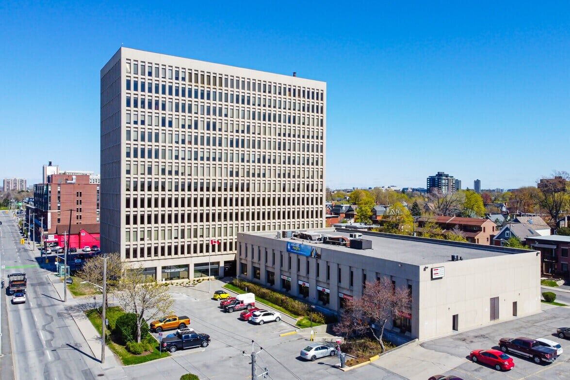 Aerial view of a cityscape with a prominent multi-story office building, smaller structures, and vehicles on a sunny day with clear skies.